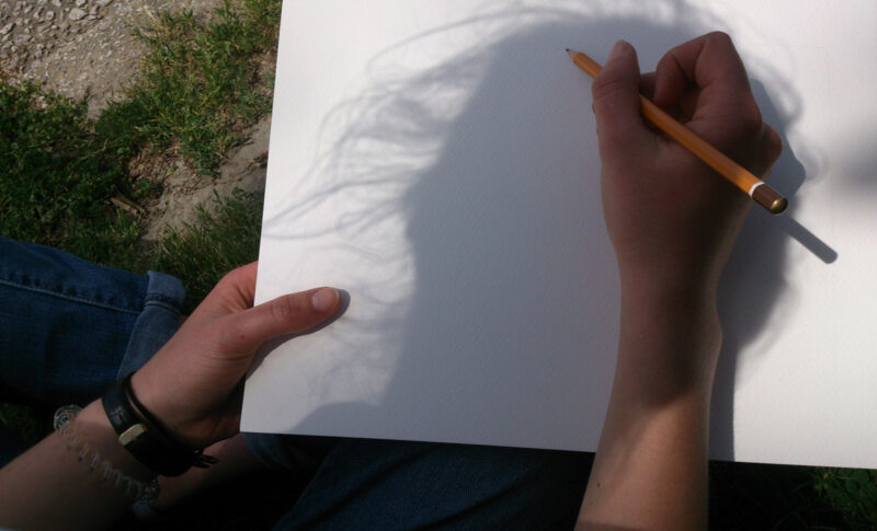L'ombre d'une tête sur une feuille de dessin, où une main s'apprête à dessiner.